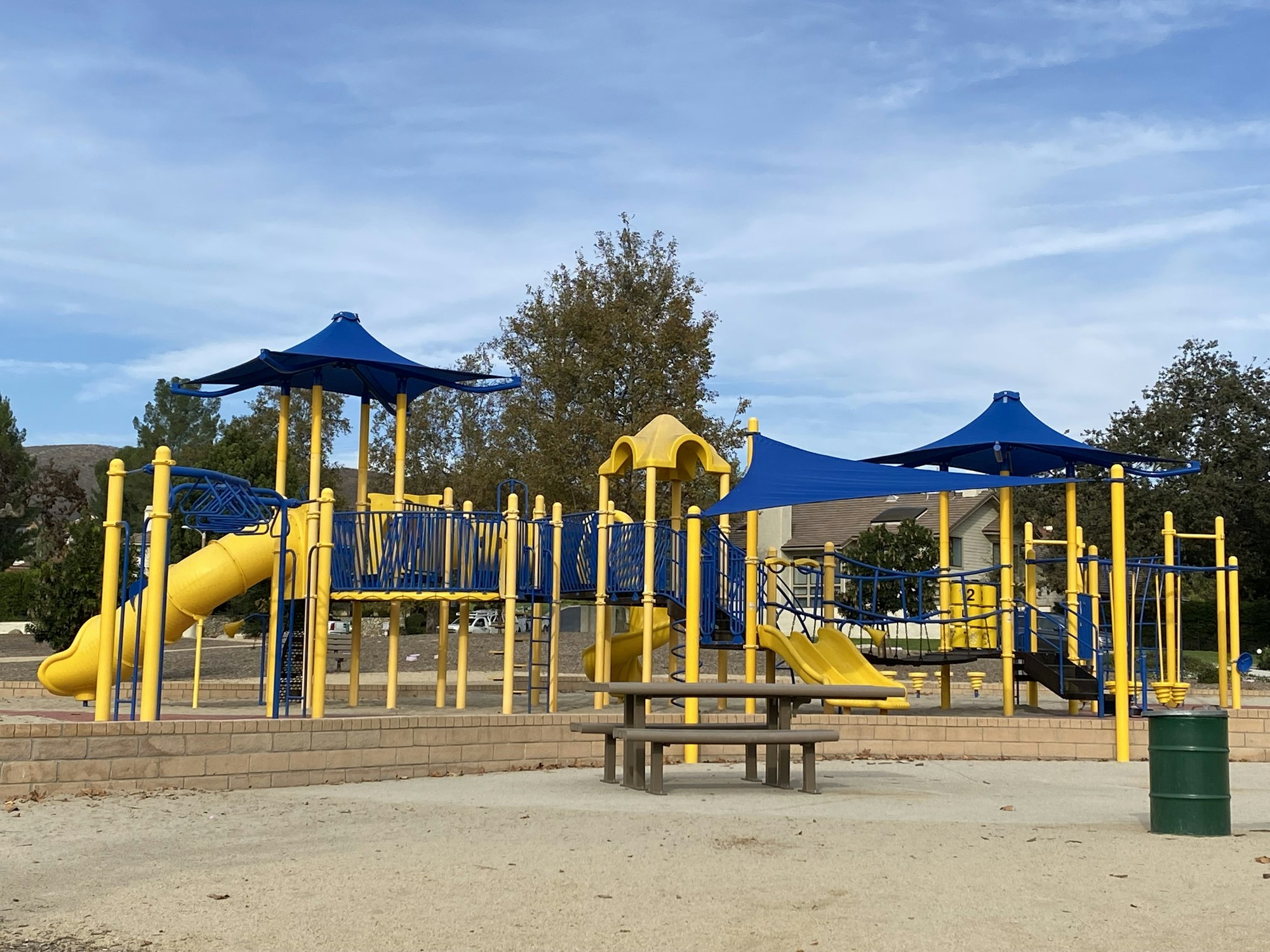 A playground with yellow slides, blue climbing structures, a picnic table, and shaded areas under a blue sky.