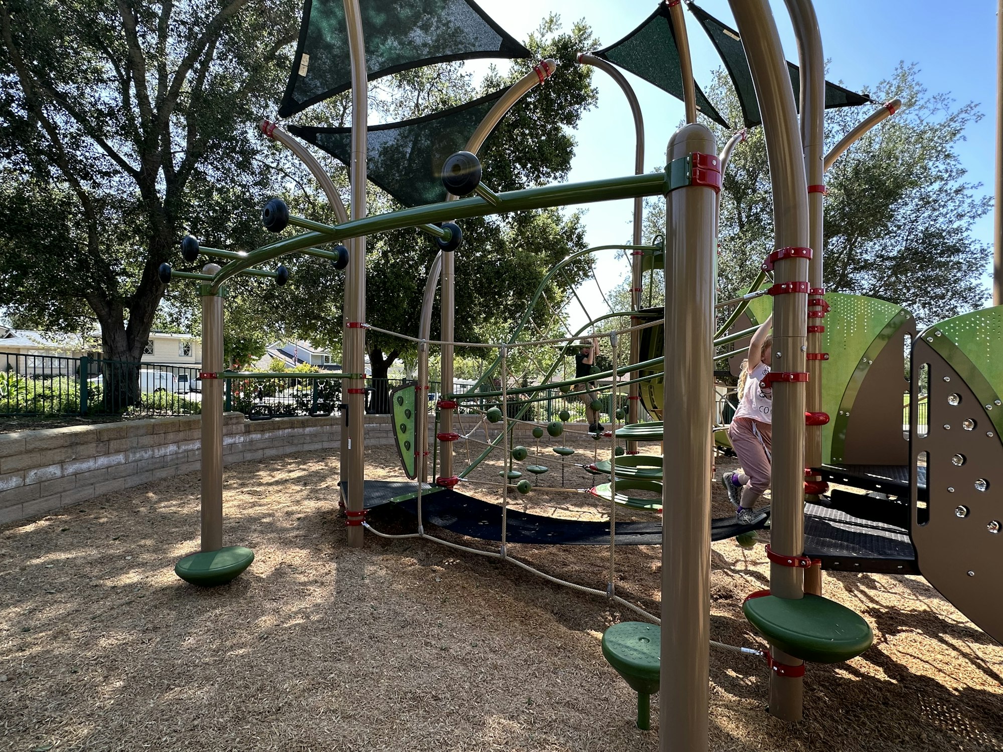 Playground with climbing structures, nets, and slides. A child is playing, surrounded by trees.