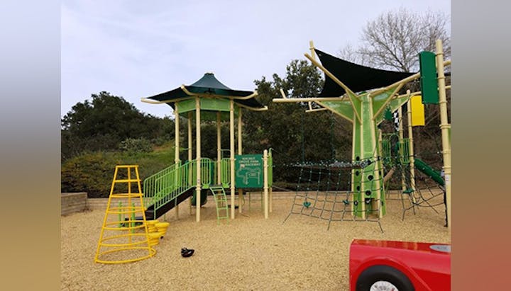 Playground with slides, climbing structures, rope nets, and sandy ground. Trees and greenery in the background.