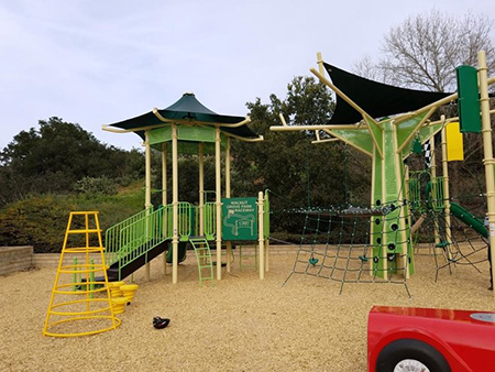 Playground with slides, climbing structures, rope nets, and sandy ground. Trees and greenery in the background.