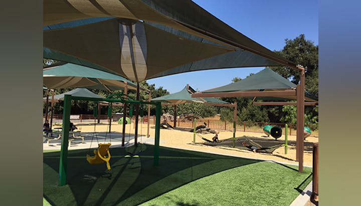 A sunny playground with shade structures, swings, picnic tables, and play equipment surrounded by trees.