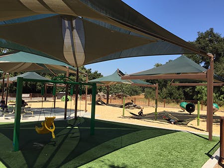 A sunny playground with shade structures, swings, picnic tables, and play equipment surrounded by trees.