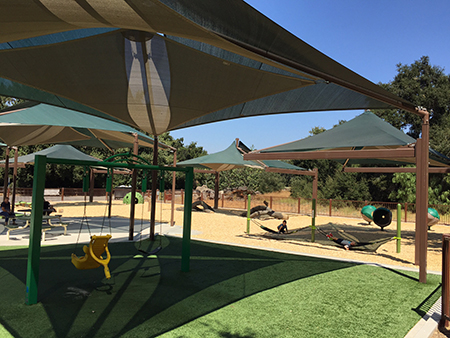 A sunny playground with shade structures, swings, picnic tables, and play equipment surrounded by trees.