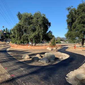 A winding blacktop path surrounded by dirt and rocks, with trees and an orange barrier in the background under a clear blue sky.