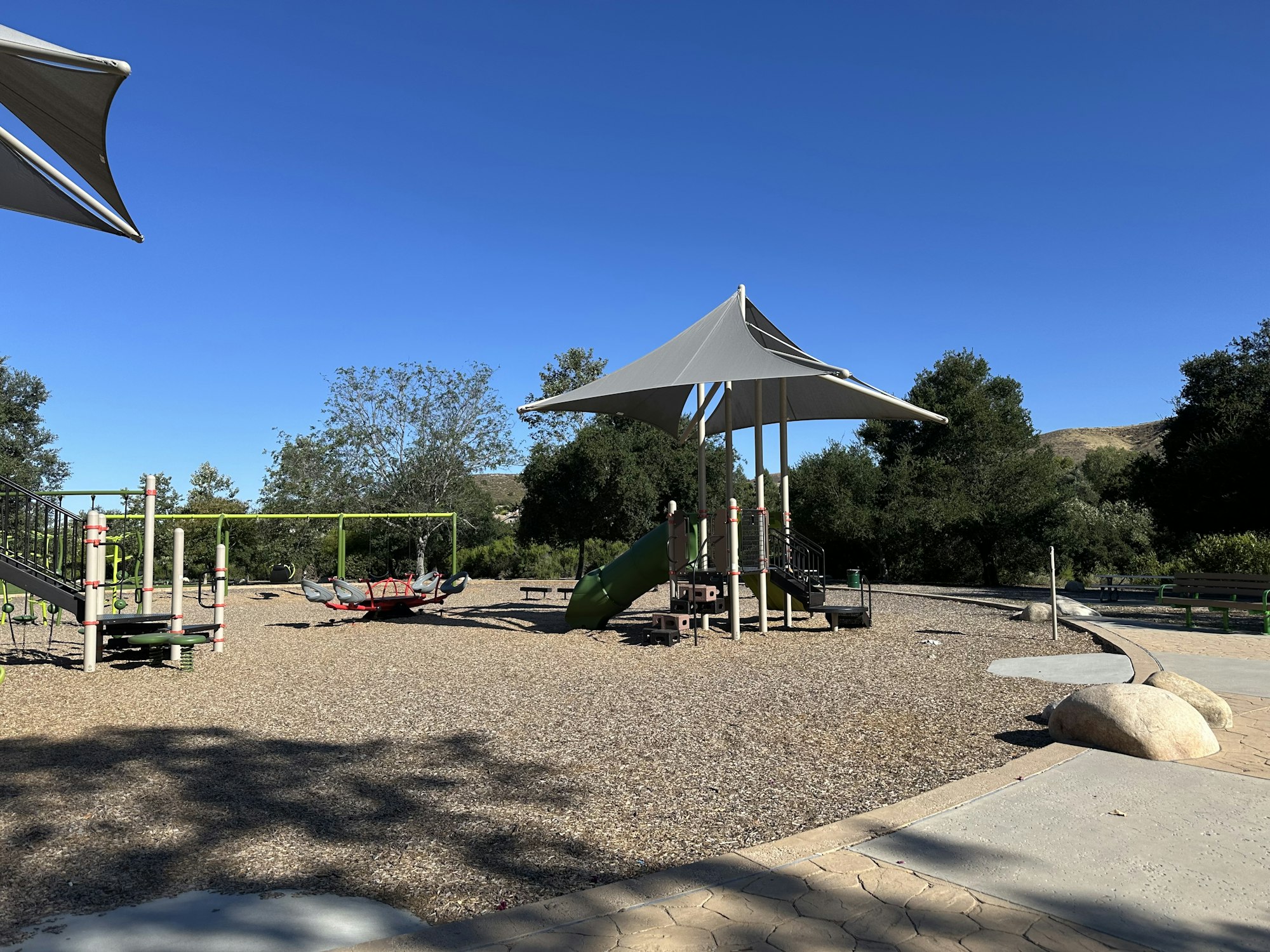 A playground scene with slide, swings, and shade structures set against a clear blue sky and greenery.