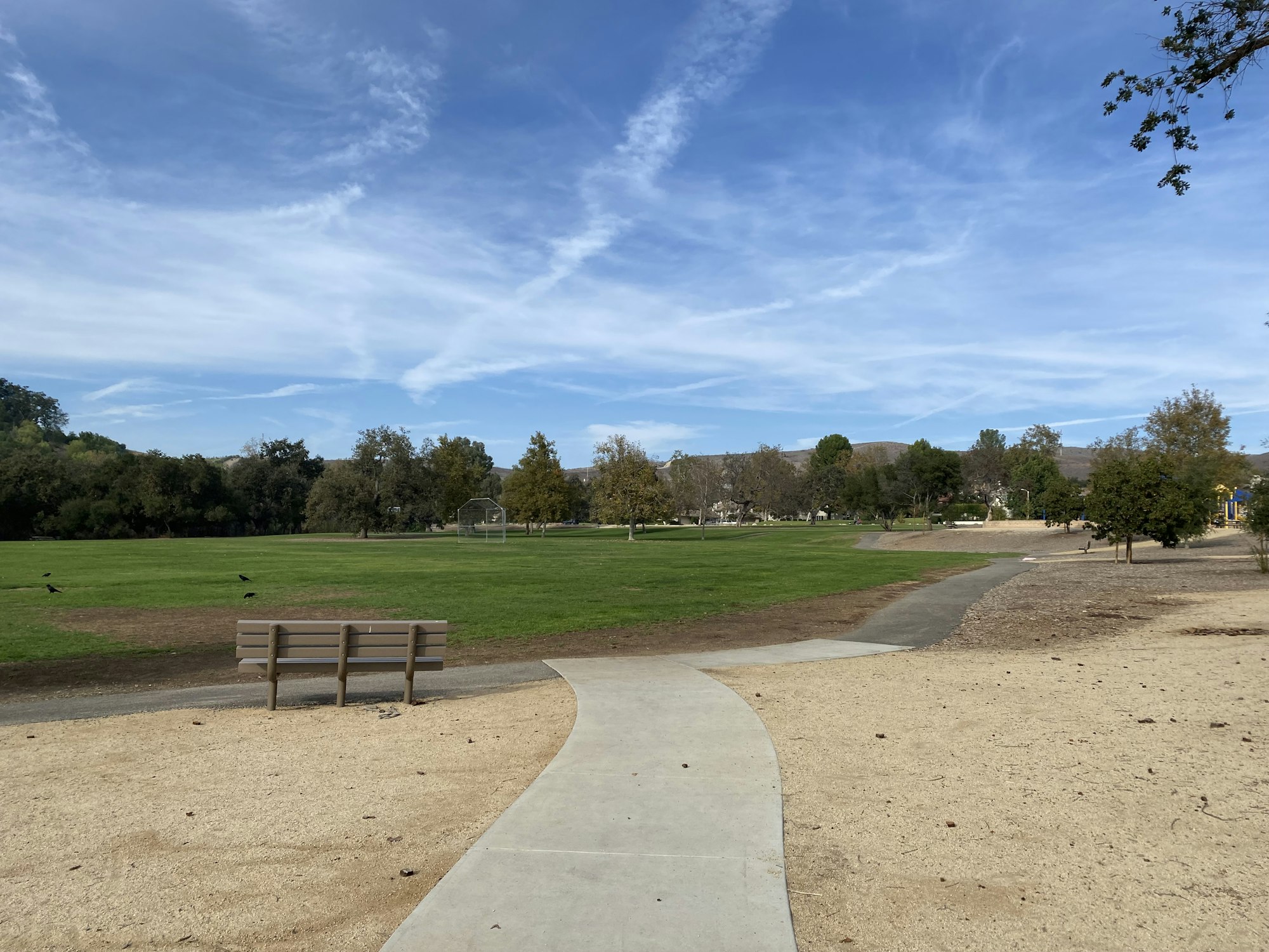 Park scene with a bench, pathway, grass field, trees, and a clear blue sky.