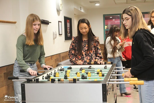 Three people playing foosball indoors, with others in the background.