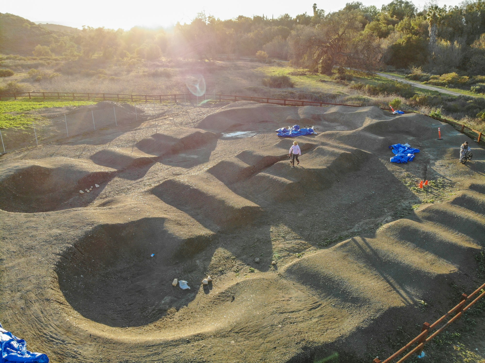 A dirt pump track with mounds, people walking, sunny outdoors setting.