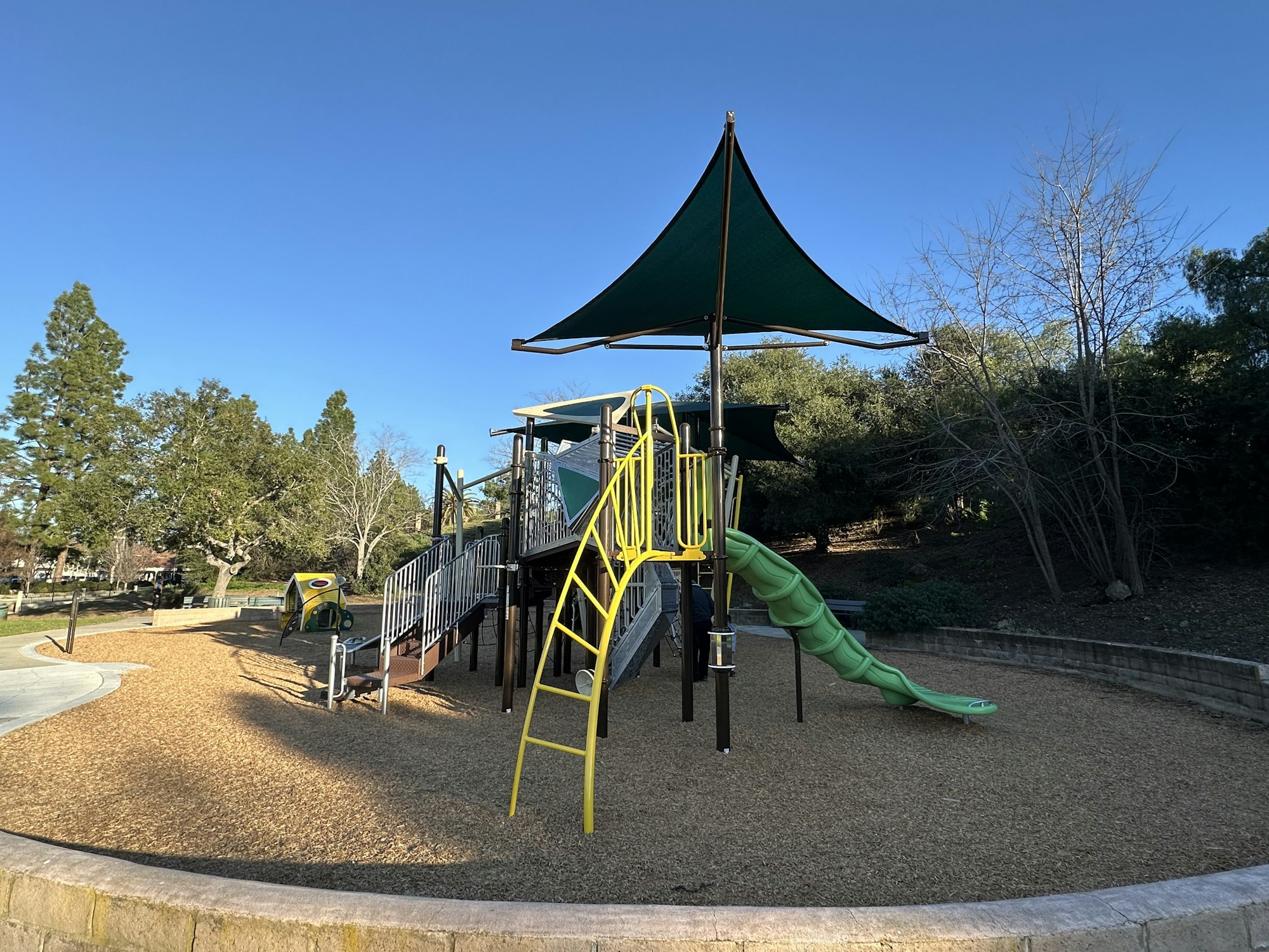 A playground with slides, climbing structures, wood chip surface, and trees in the background under a clear blue sky.