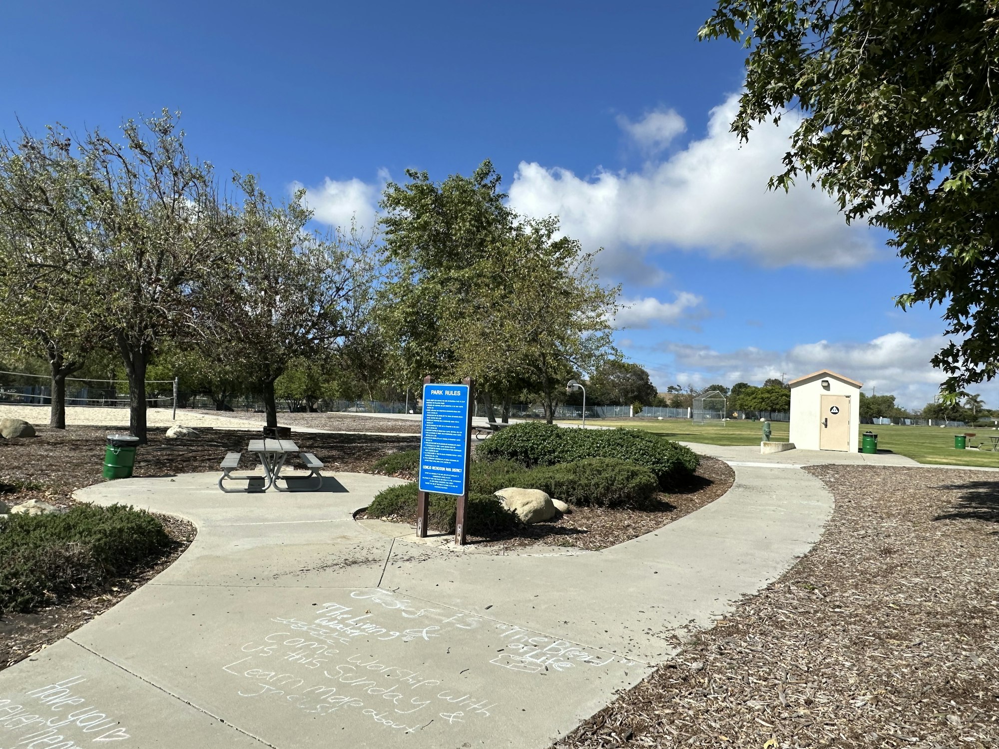 A park scene featuring picnic tables, trees, a rules sign, and a restroom building under a partly cloudy sky.