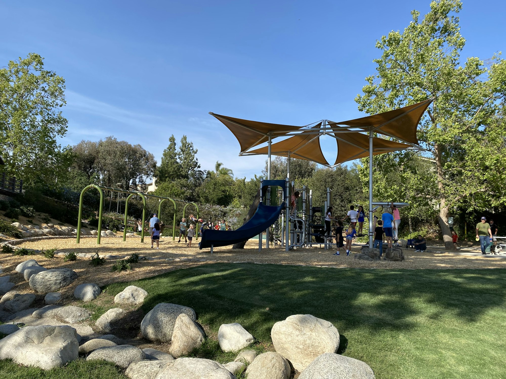 Playground with swings, a slide, kids playing, and adults watching on a sunny day surrounded by trees and rocks.