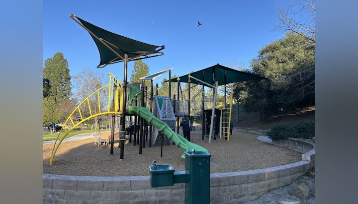 Playground with slides, climbing structures, shade canopies, a drinking fountain, surrounded by trees and wood chips.