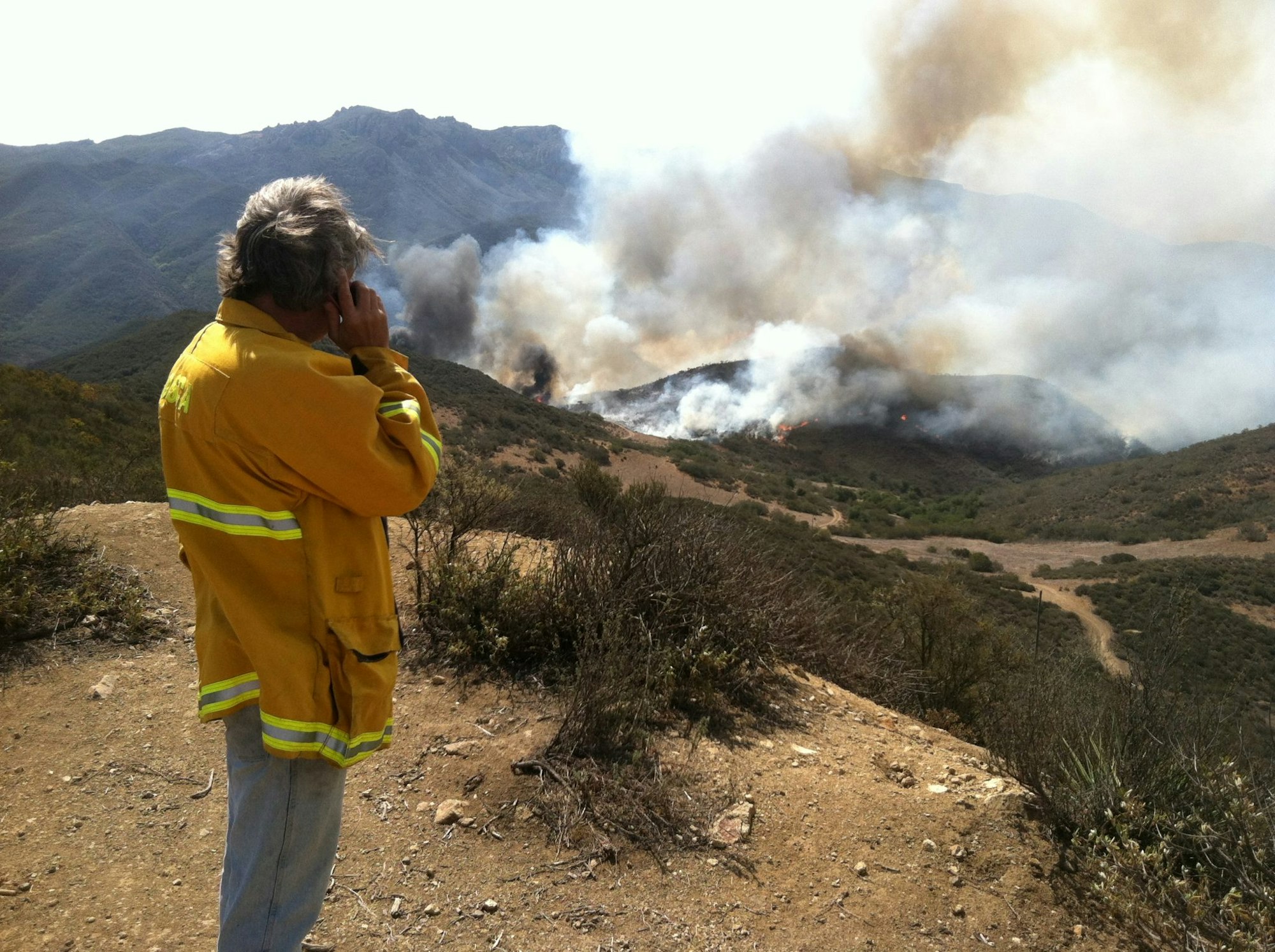 A person in a yellow jacket is talking on a phone, observing smoke and fire in a hilly landscape.