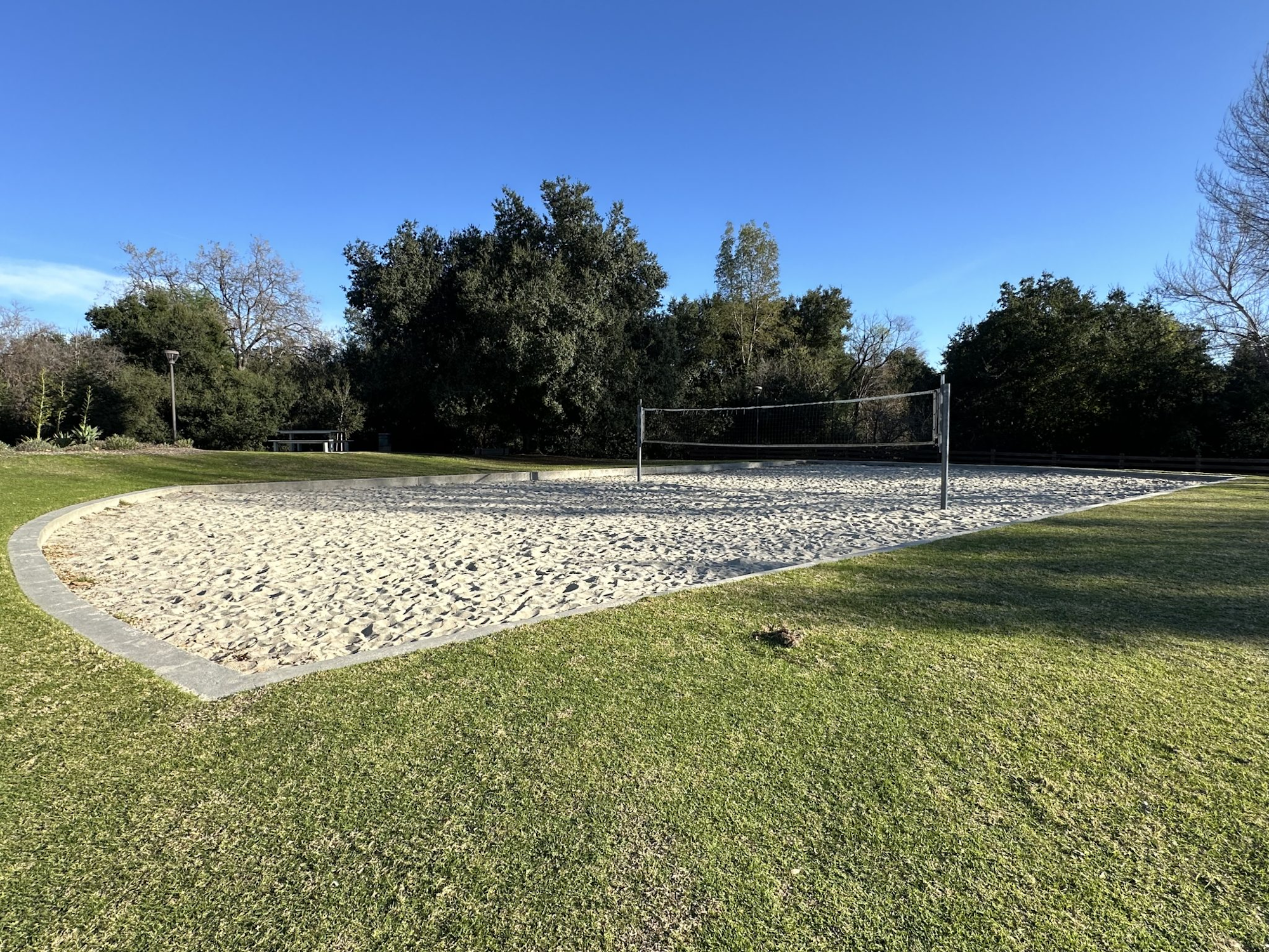 A sand volleyball court surrounded by grass and trees under a clear blue sky.