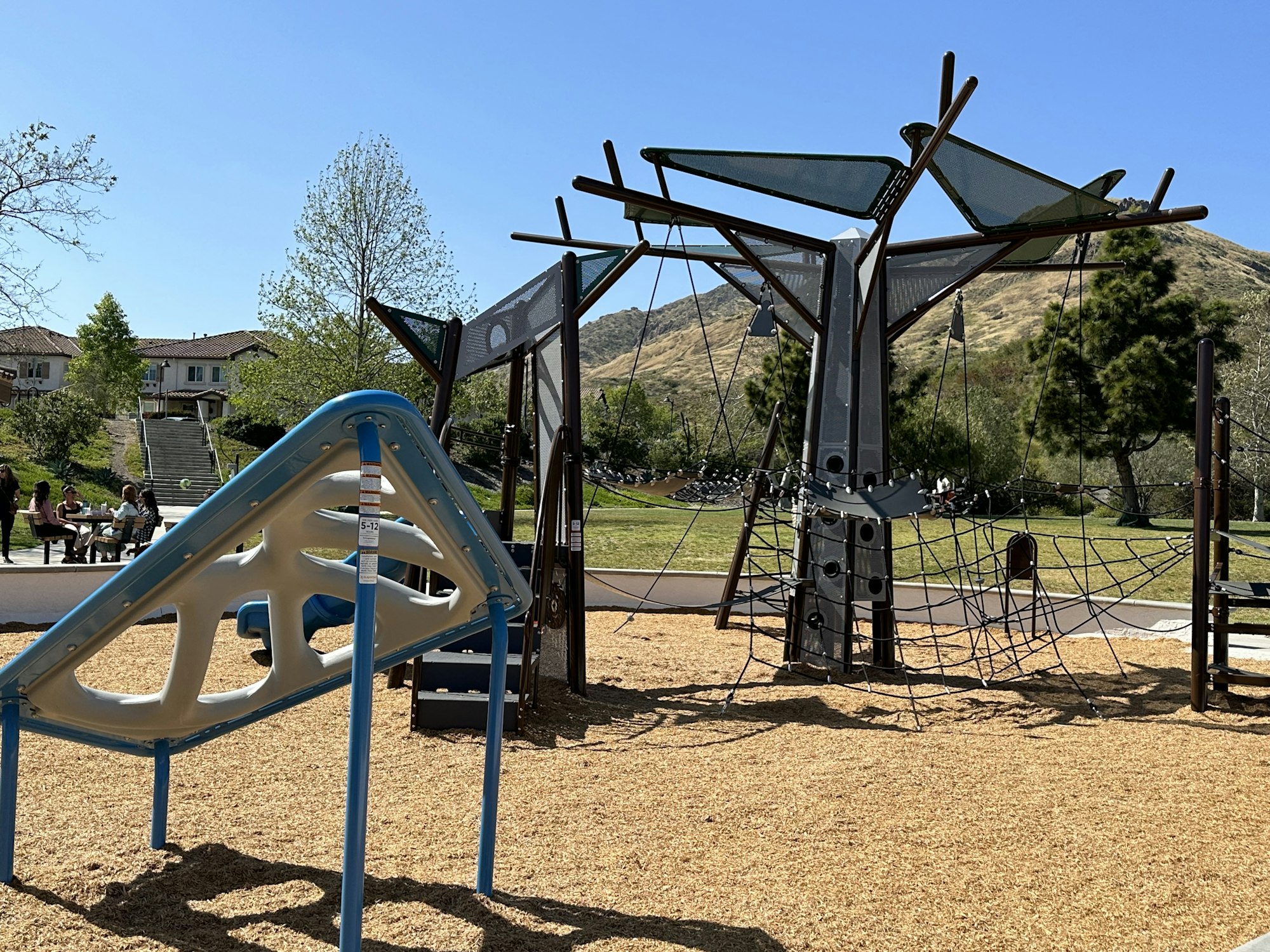 A playground with climbing structures, ropes, and slides. There are people sitting at a picnic table nearby, with trees and hills in the background.