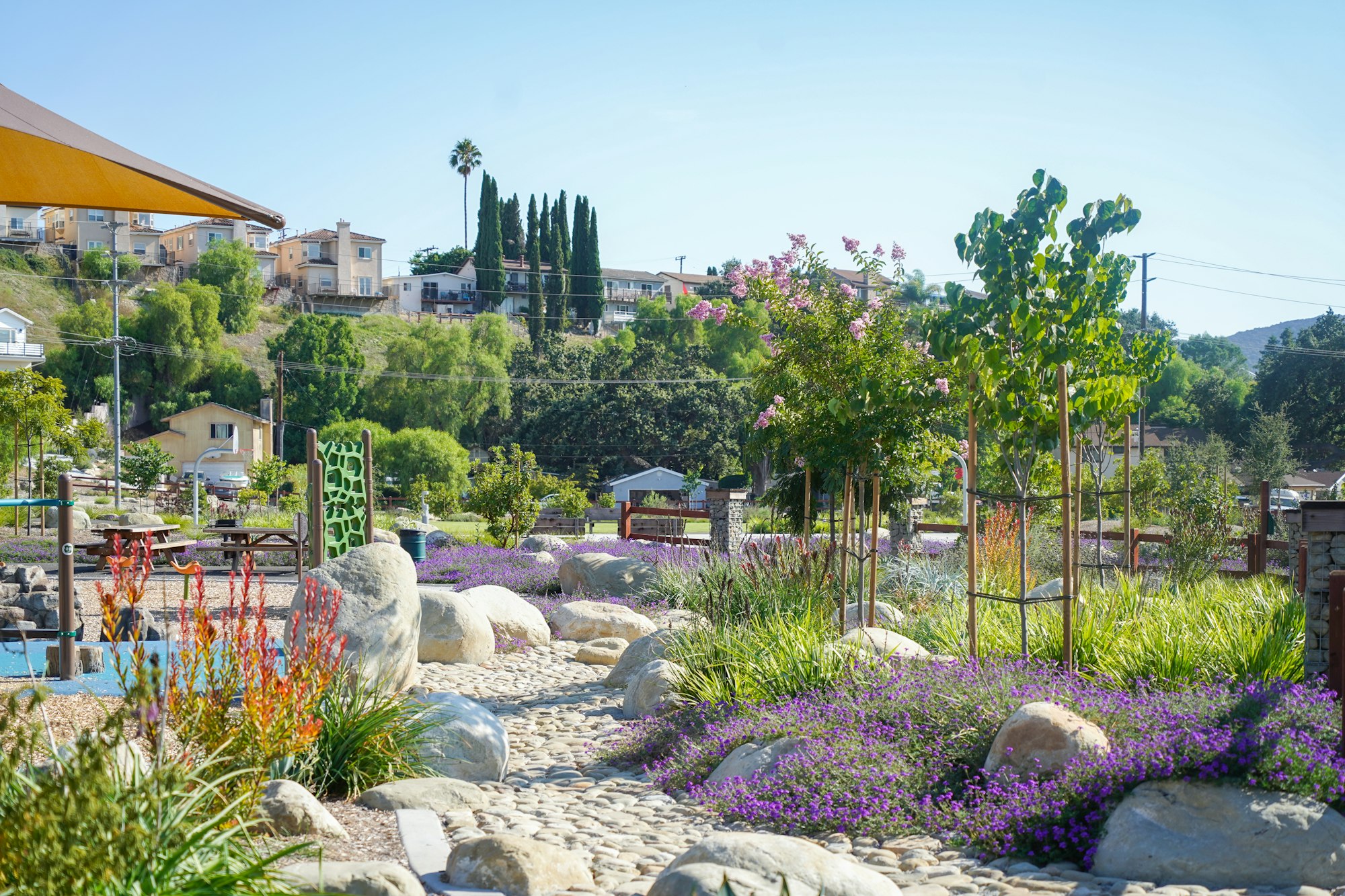 A landscaped garden with rocks, flowers, trees, and a path. Houses and hills are visible in the background.