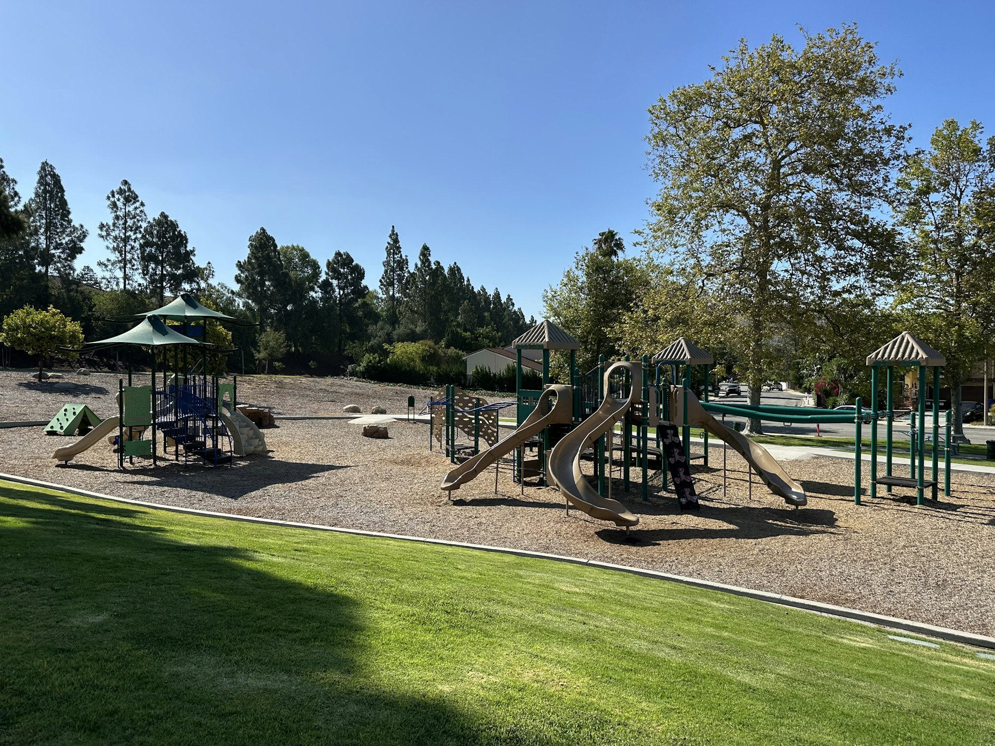 A sunny playground featuring slides, climbing structures, and grassy areas surrounded by trees.