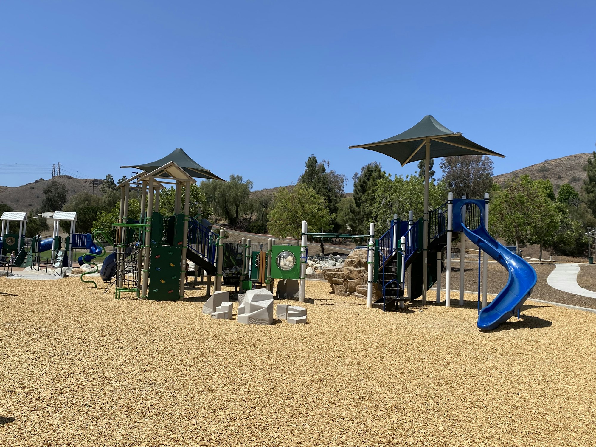 Playground with slides, climbing structures, and shade canopies on mulch ground, surrounded by trees and a clear blue sky.