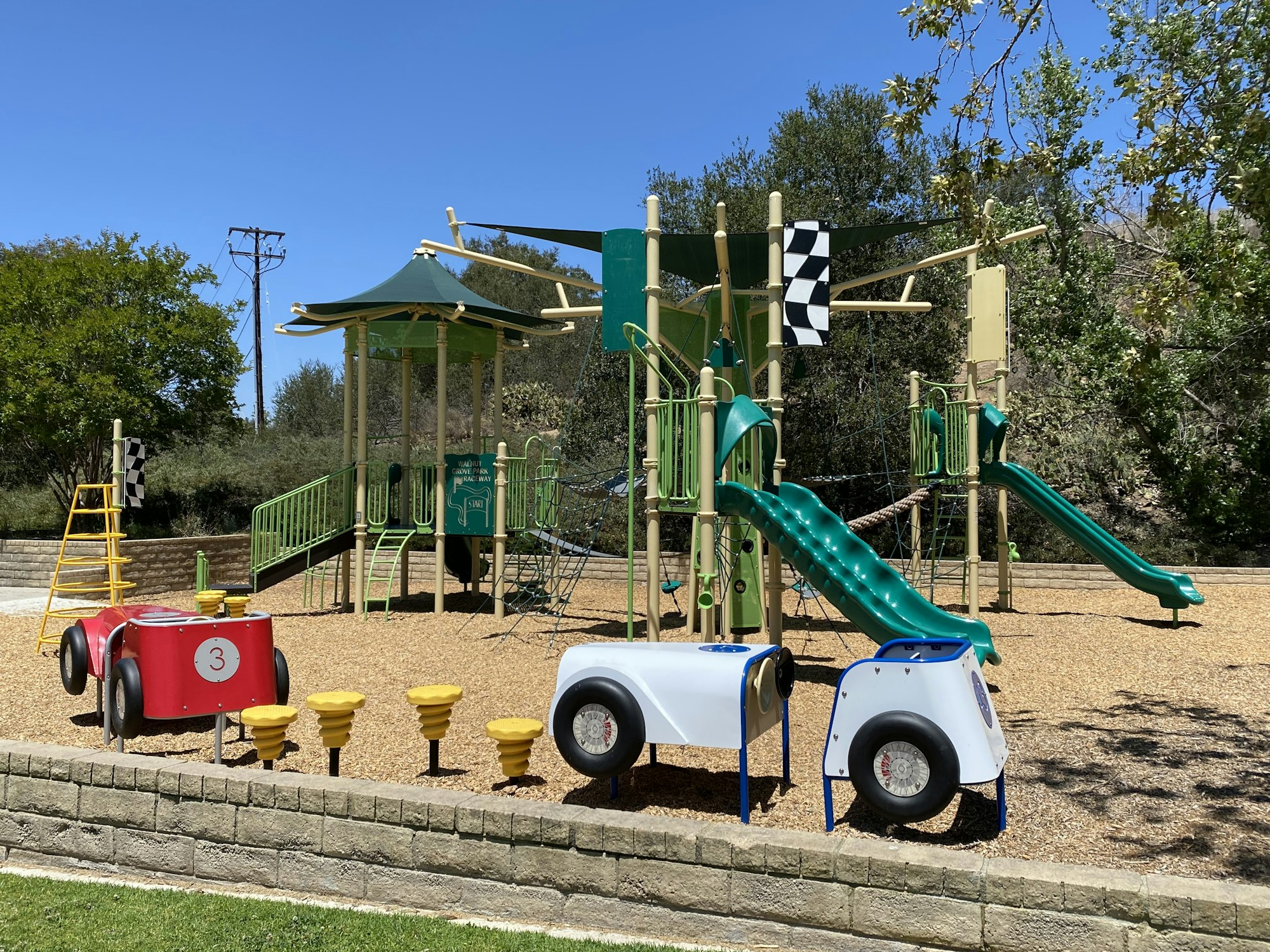 Children's playground with green slides, climbing structures, a red and white car structure, and a checkered flag.