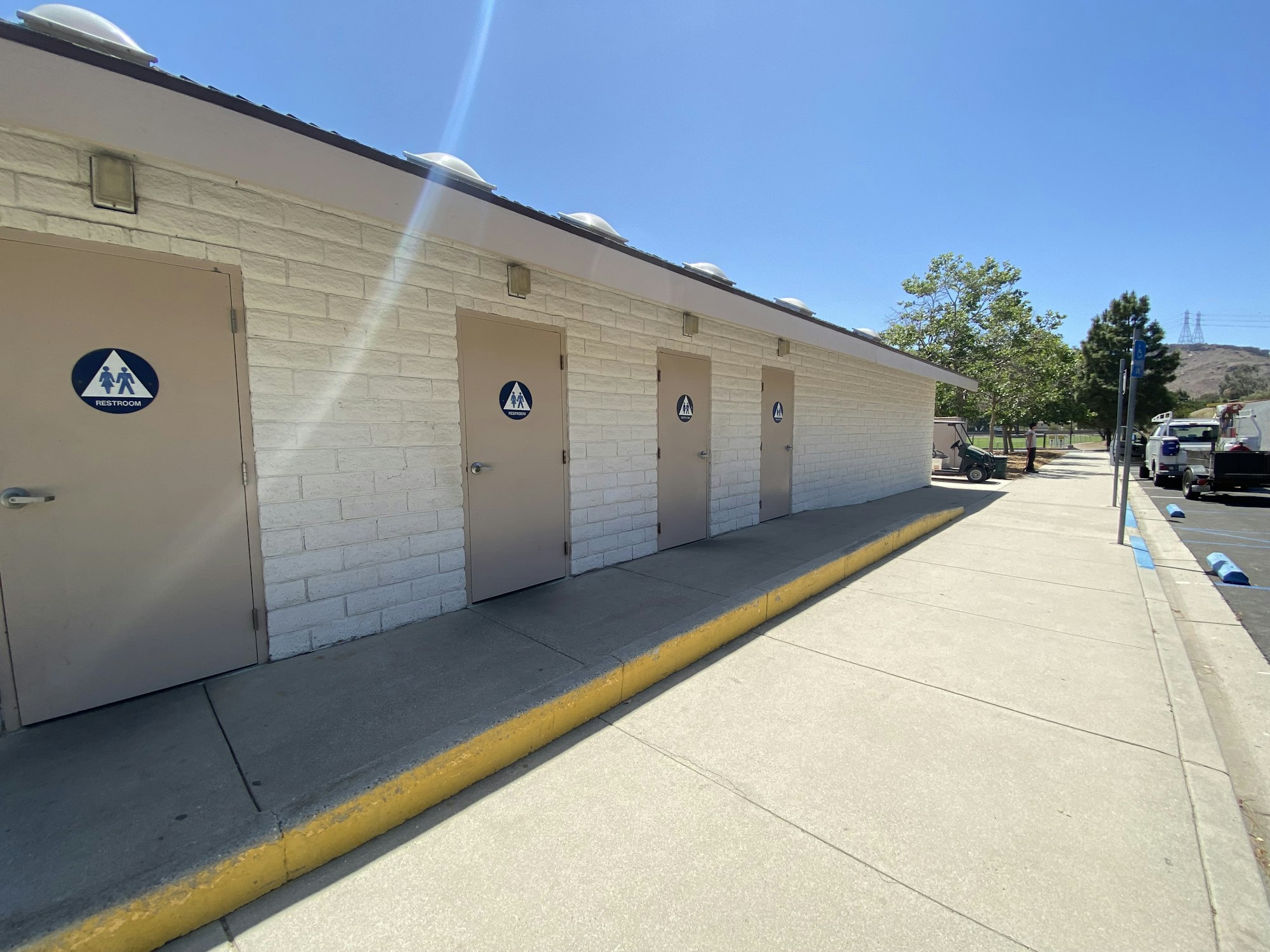 A row of restroom doors outside a building under a clear sky, with a sidewalk and parked vehicles nearby.