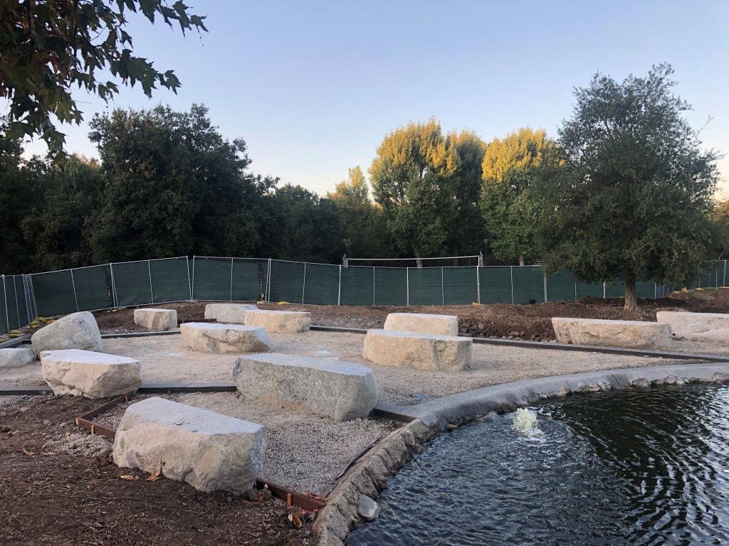 Large rocks on gravel by a pond, fenced area, and trees in the background.