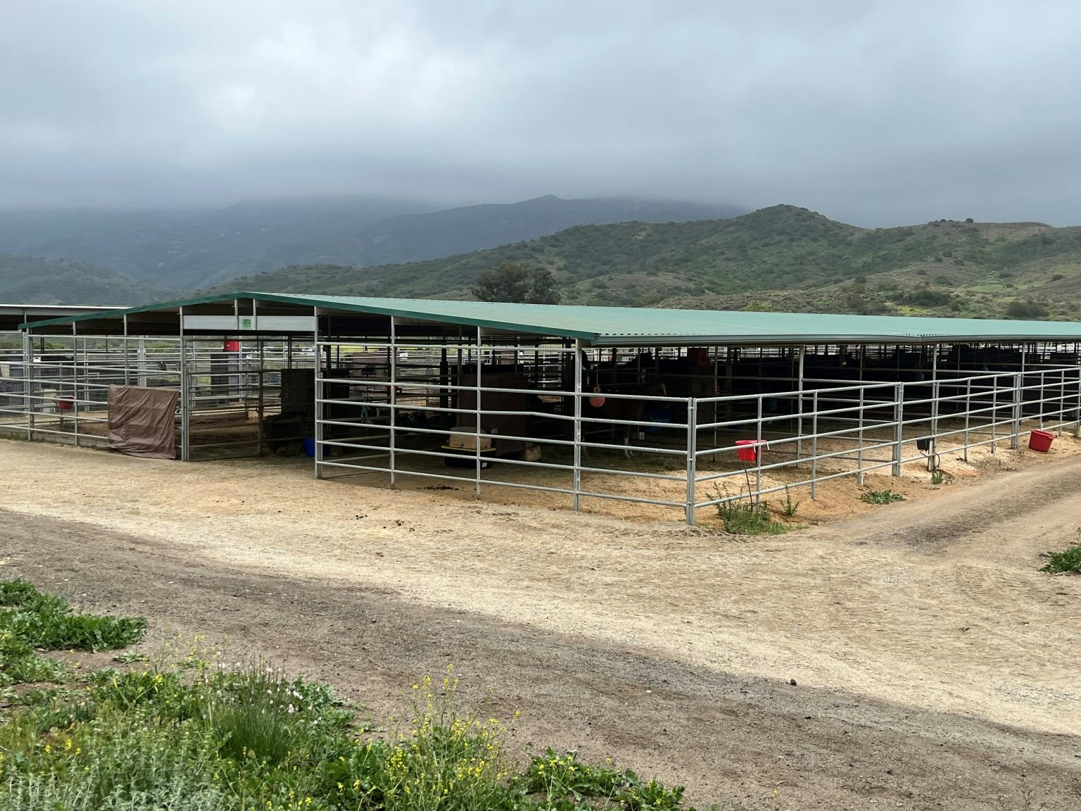 A fenced area with metal roofs, possibly horse stables, on a cloudy day with green hills in the background.