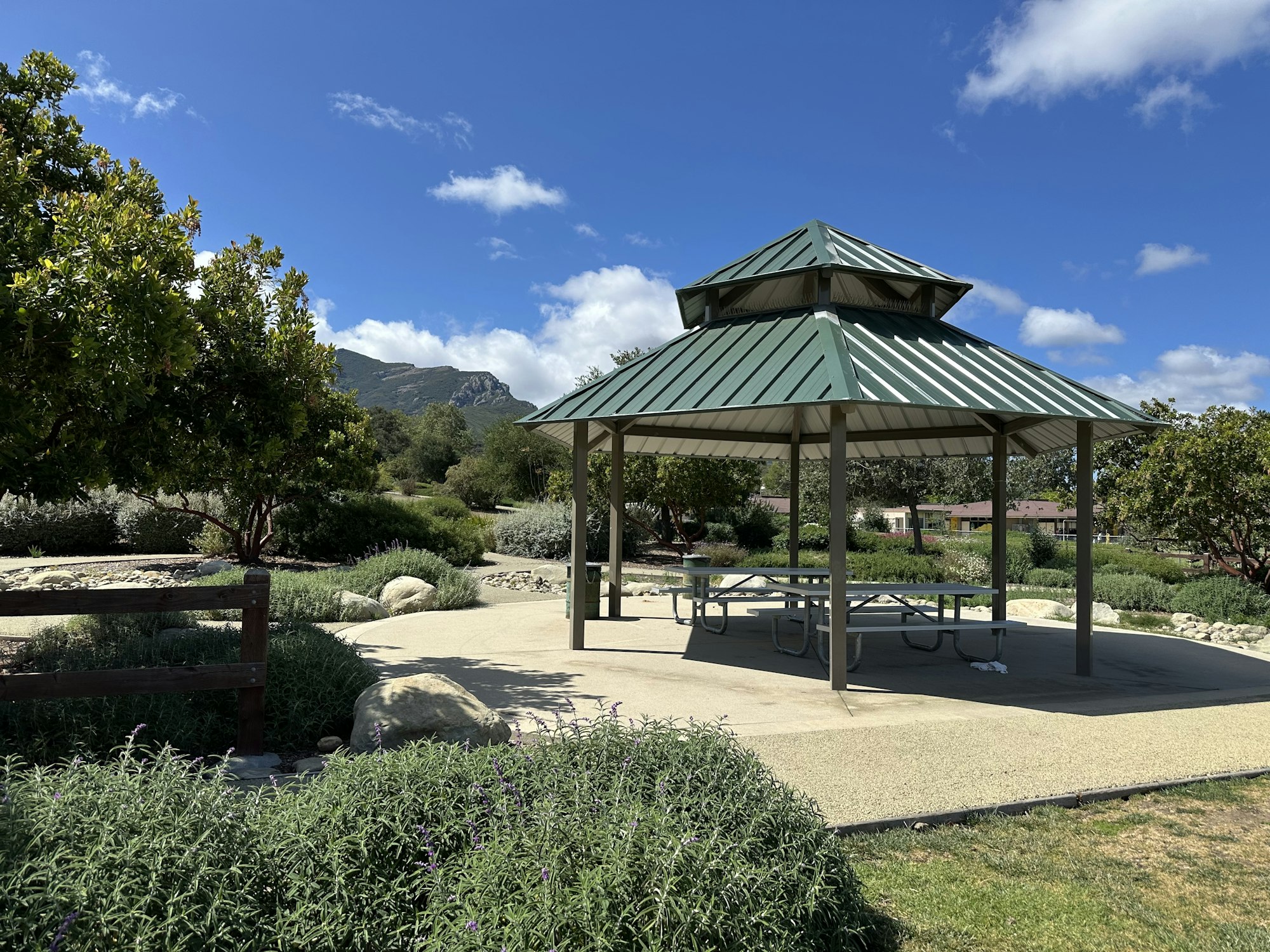 A park gazebo with a green roof, surrounded by greenery and rocks, against a backdrop of mountains and a blue sky.