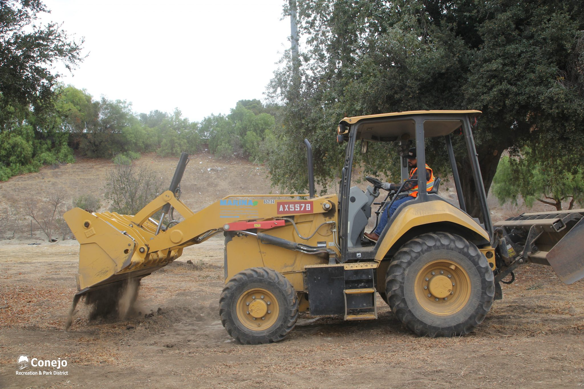 A person operates a yellow backhoe loader on dirt, surrounded by trees.