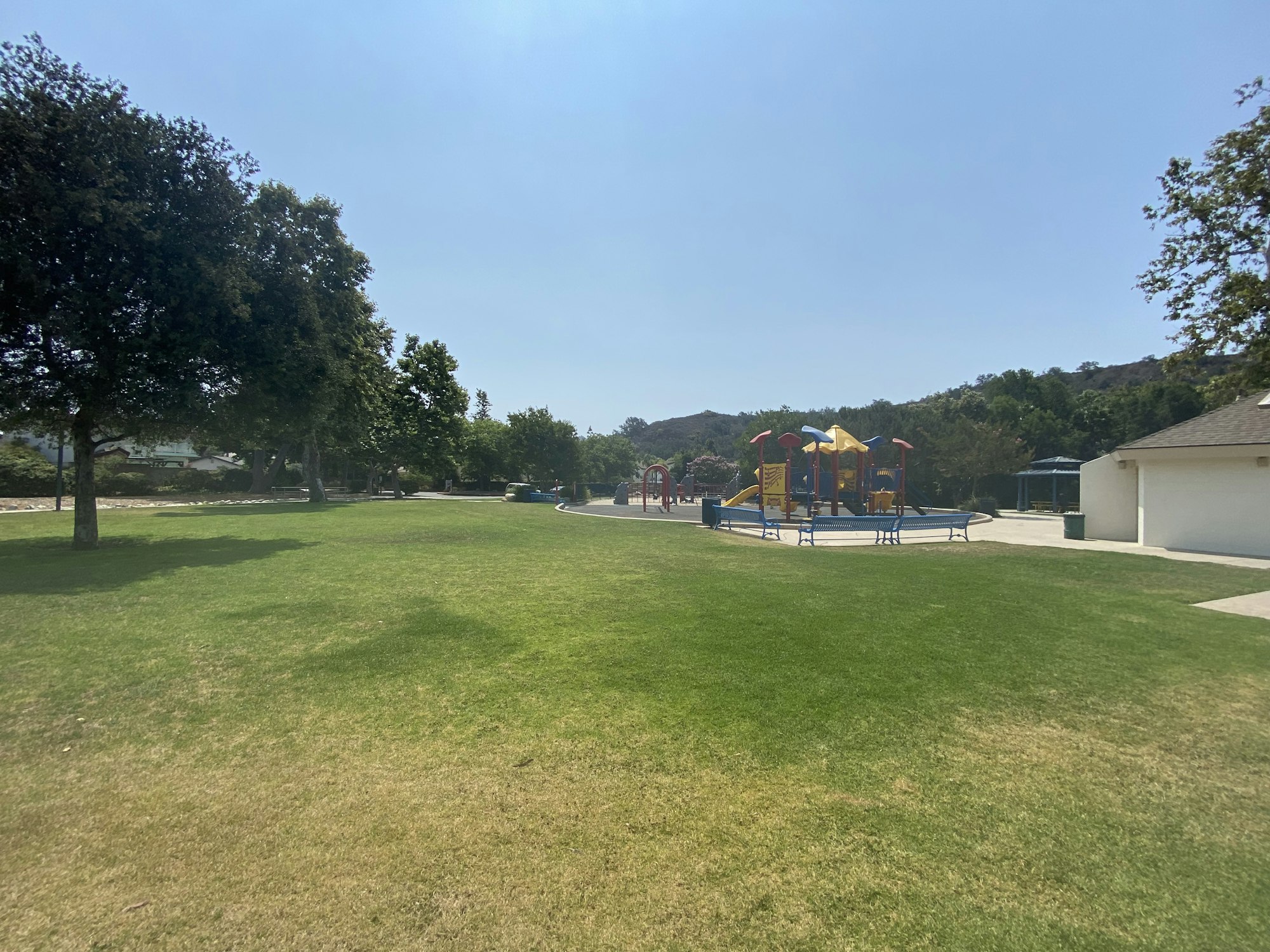 A grassy park with trees, a play structure, and a building under a clear sky.