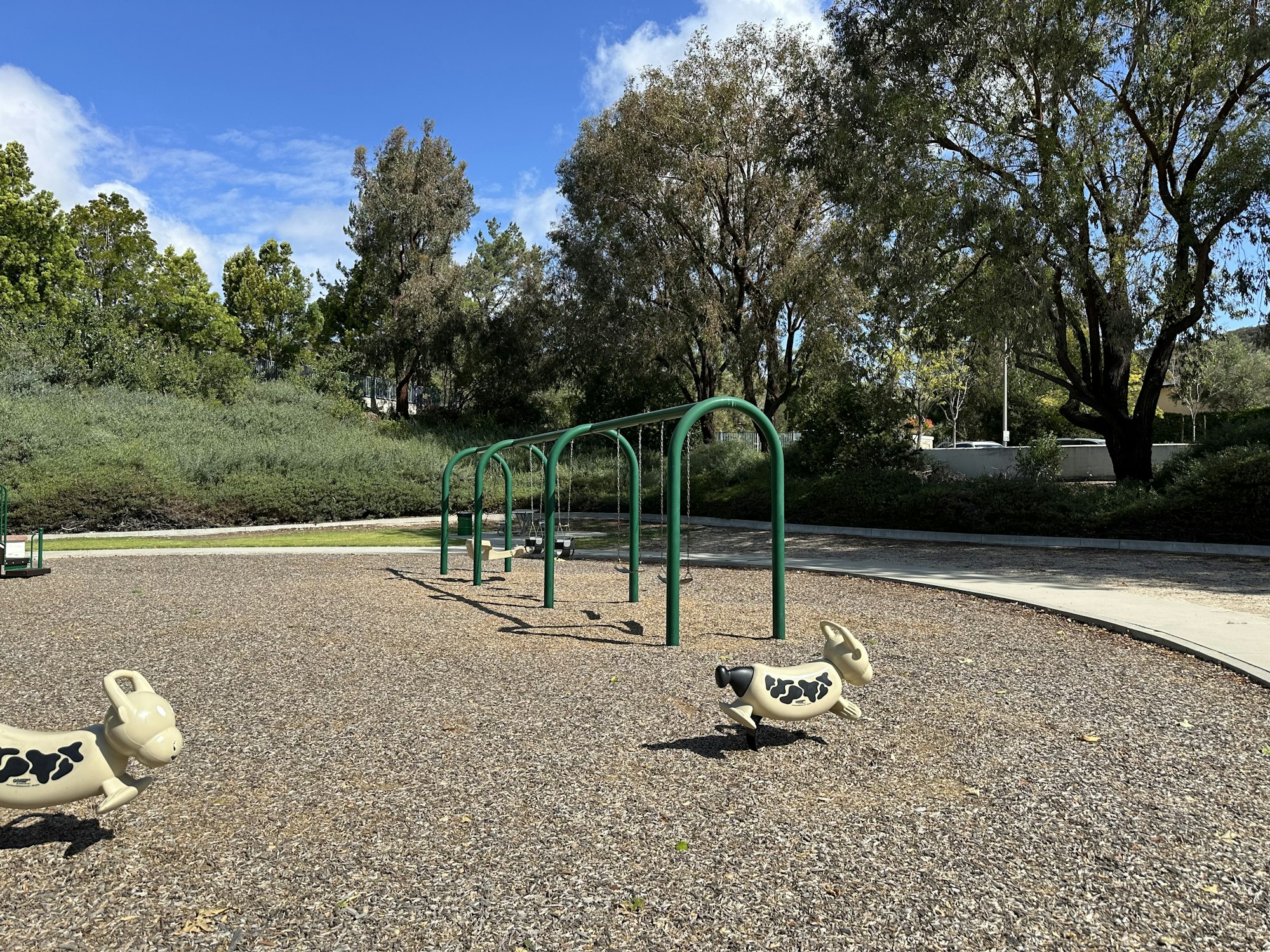 An empty playground featuring swings and two cow-shaped spring riders, surrounded by trees and blue skies.