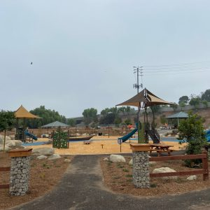A playground with slides, climbing structures, and paths, surrounded by trees and a fence.