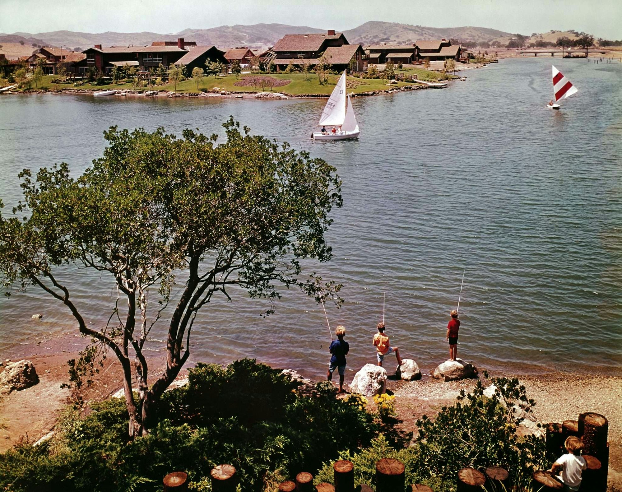 Children fishing by a lake with sailboats, houses, and hills in the background.
