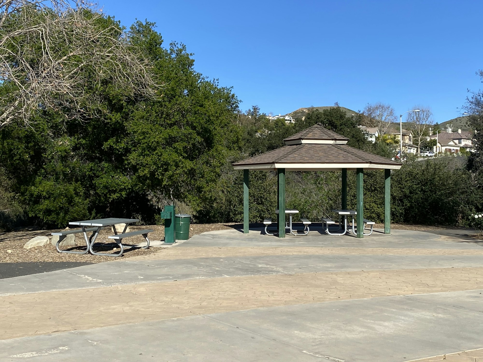 A park scene with a wooden pavilion, picnic tables, trash cans, and trees surrounding a path.