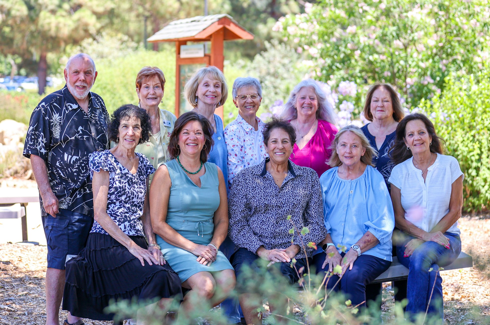 A group of smiling people posing outdoors with greenery in the background.