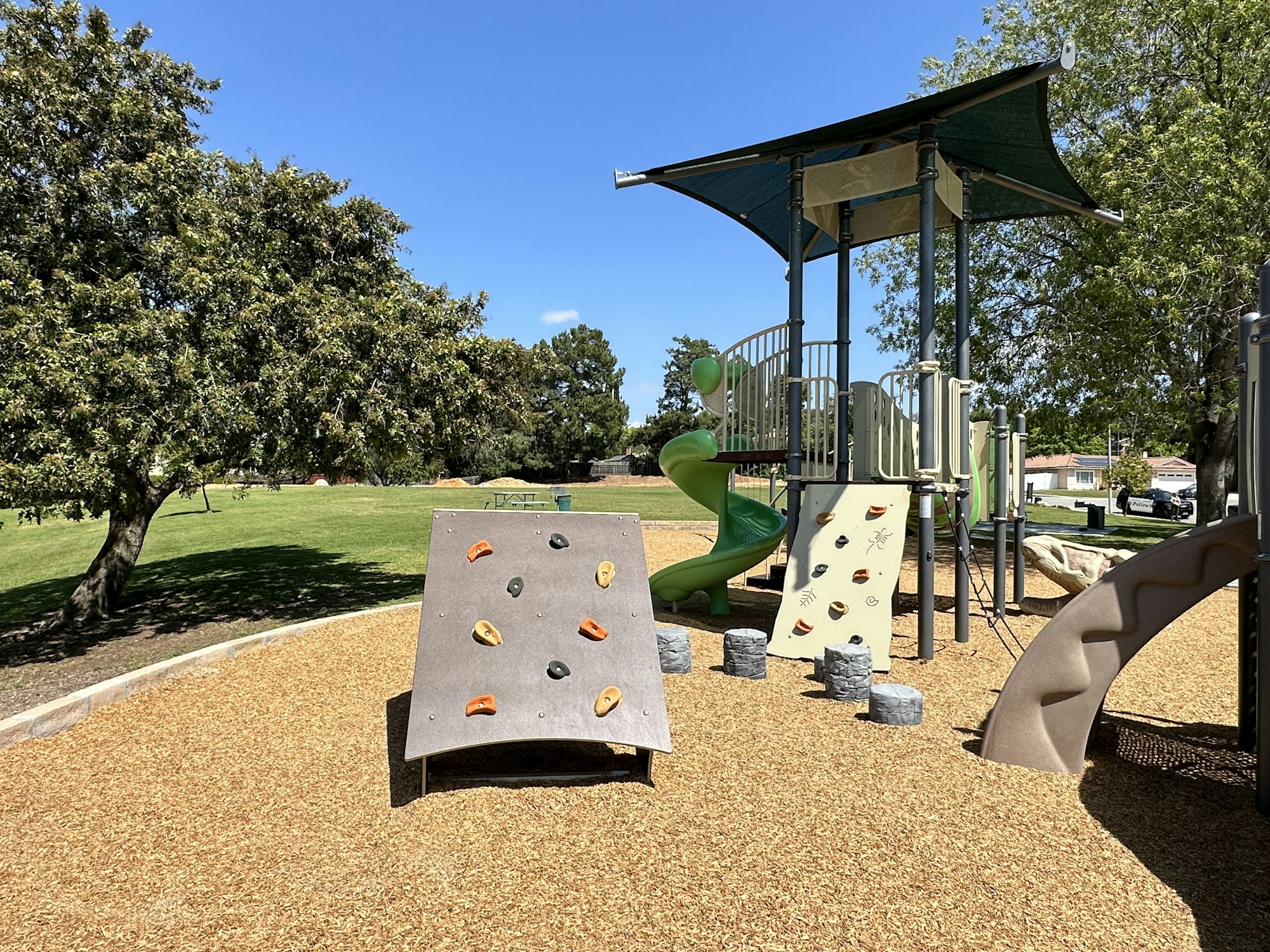 A playground with climbing structures, a slide, and artificial flooring, surrounded by trees and a grassy field under a clear blue sky.