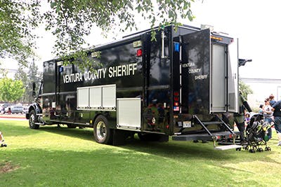 A large black Ventura County Sheriff vehicle is parked on grass, with people and a stroller nearby.