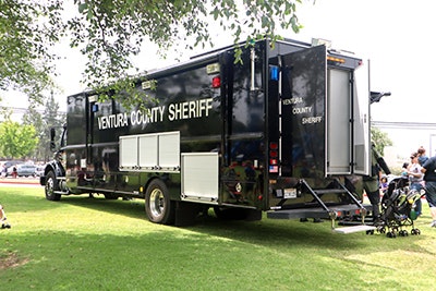 A large black Ventura County Sheriff vehicle is parked on grass, with people and a stroller nearby.
