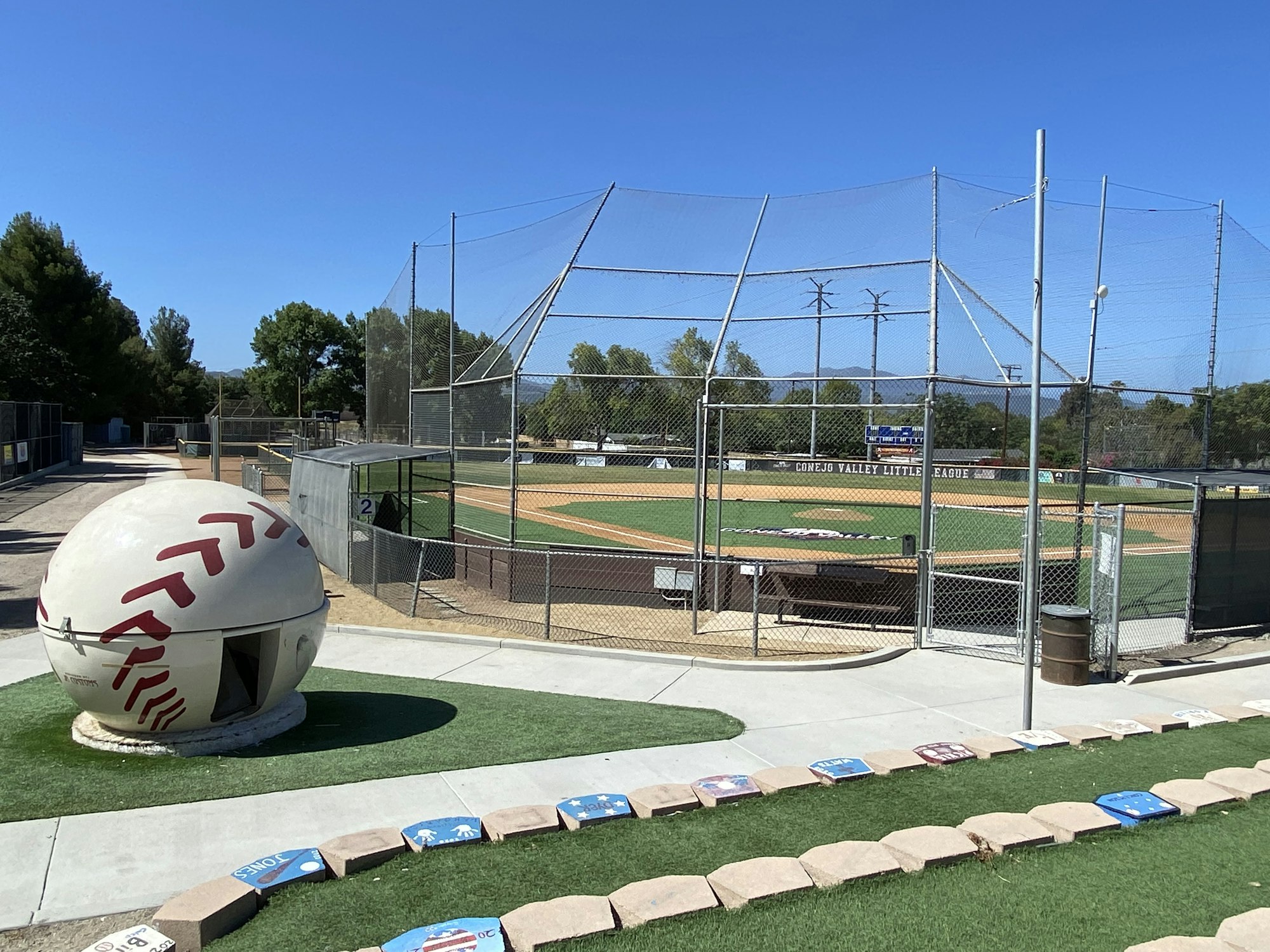 A baseball field with a large baseball sculpture nearby, surrounded by a chain-link fence and clear blue sky.