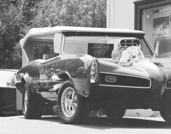 A vintage customized car with an exposed engine, "Hands Off" sign on the windshield, parked outdoors.