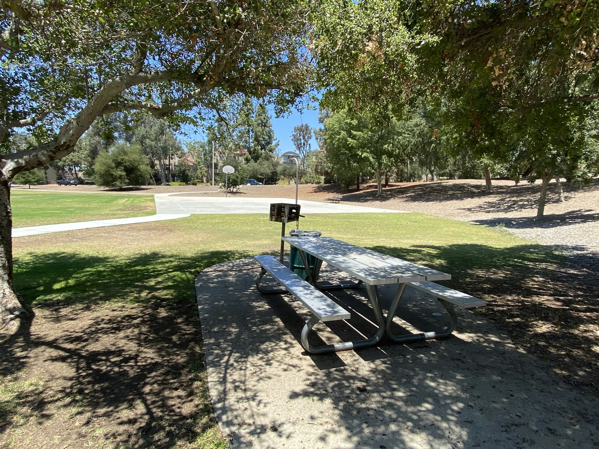 A picnic table in a sunlit park with trees and a basketball court in the background.