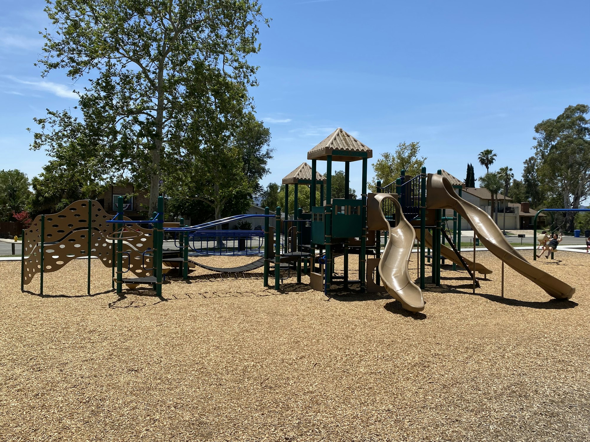 A playground with slides, climbing structures, and wood chip ground cover under a clear blue sky.