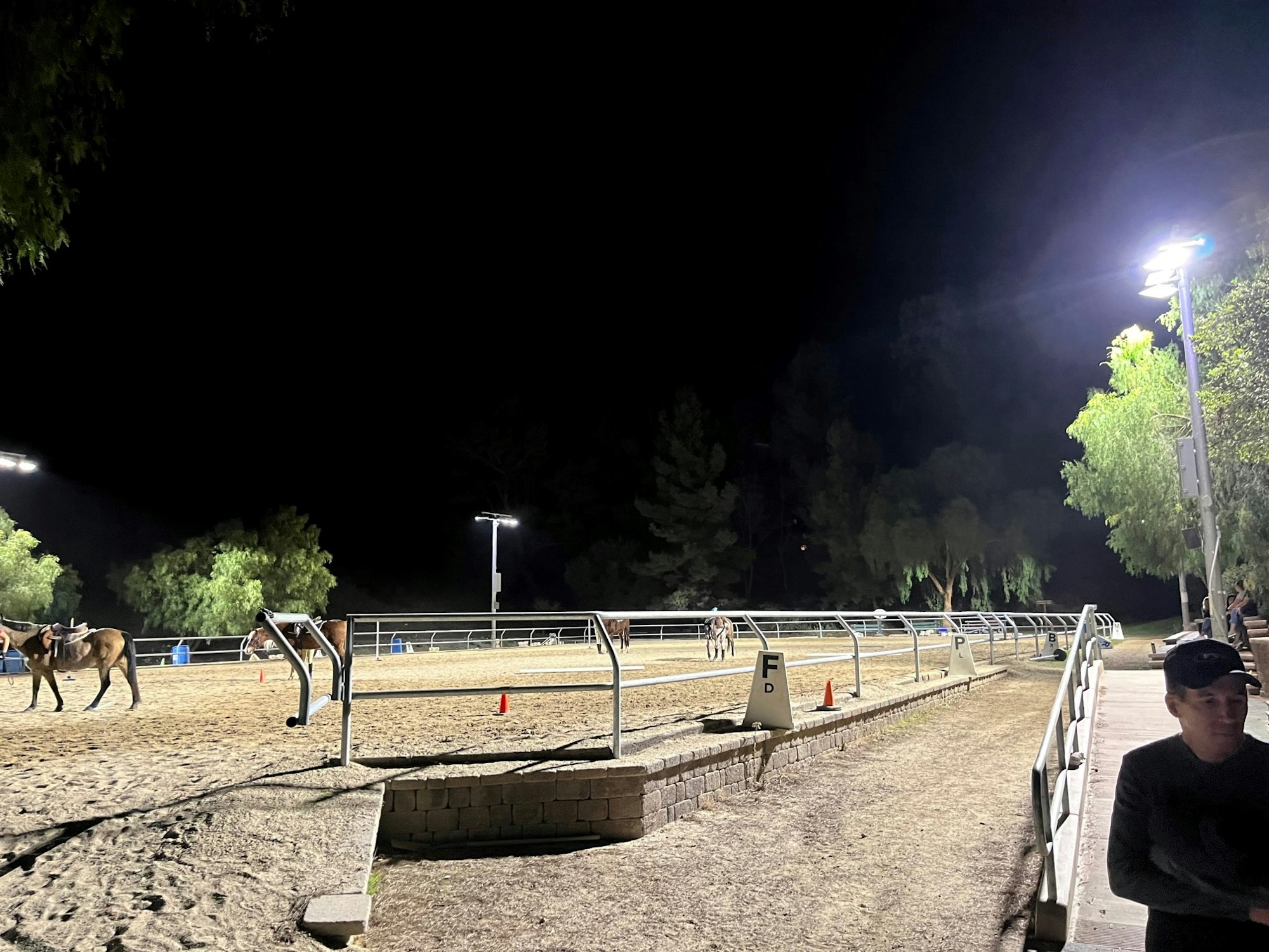 A nighttime equestrian arena, with horses and riders, illuminated by floodlights, and a person standing nearby.