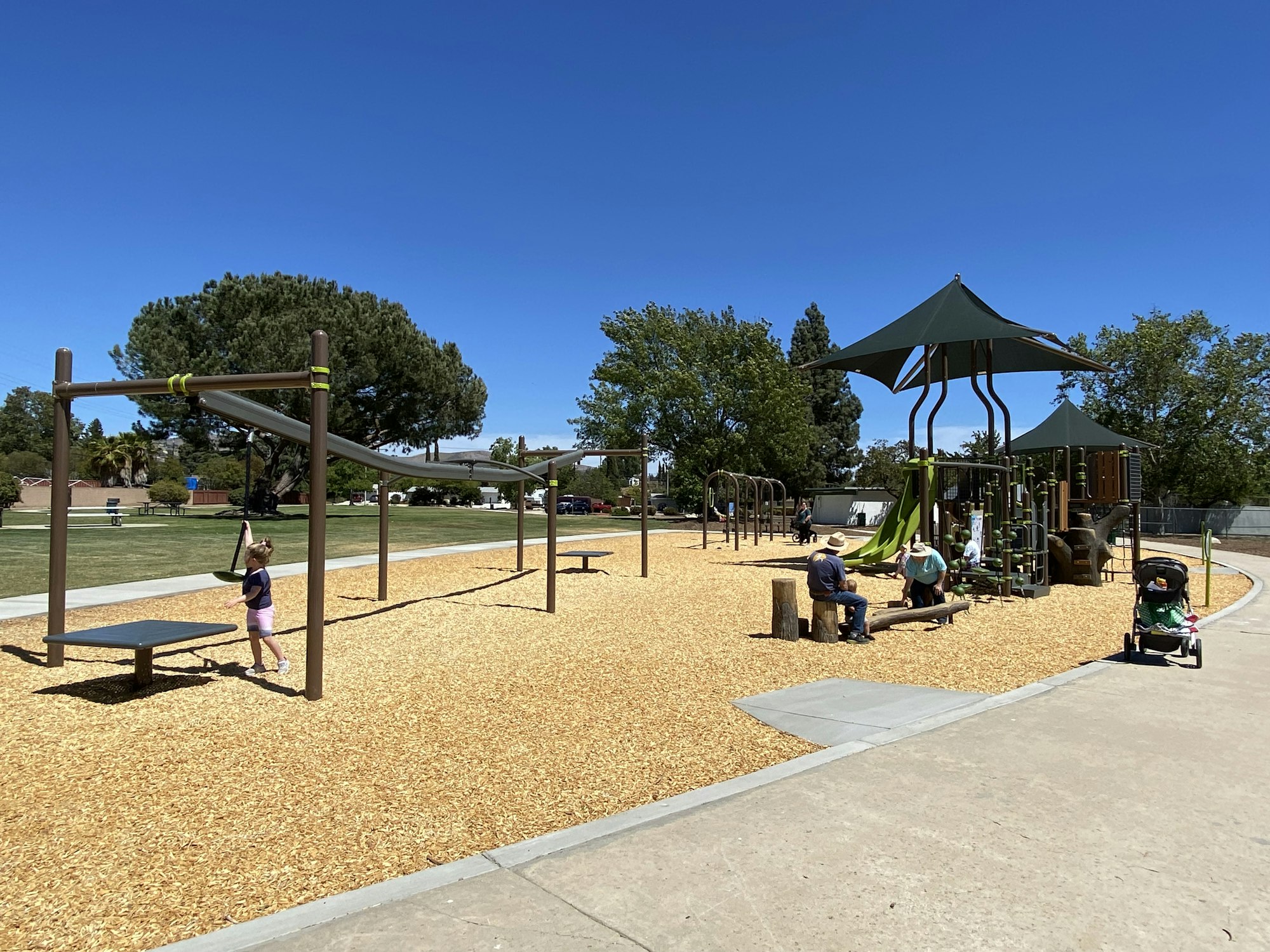 Playground with a climbing structure, a slide, benches, people, and a stroller on a sunny day.