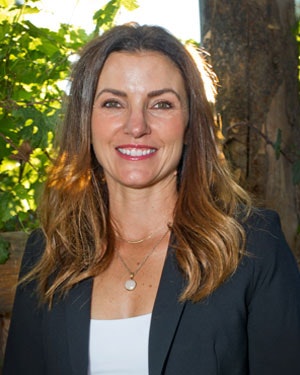 A person with long brown hair smiling, wearing a black blazer and white top, standing outdoors with greenery in the background.