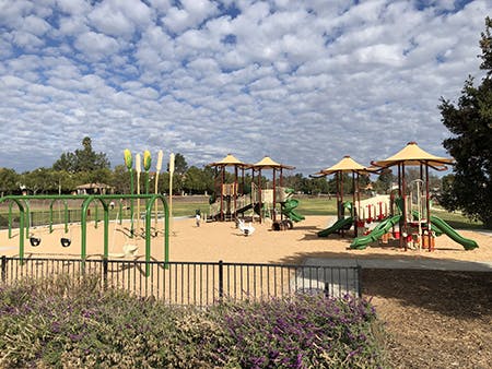 Playground with slides, swings, and climbing structures; cloudy sky and trees in the background.