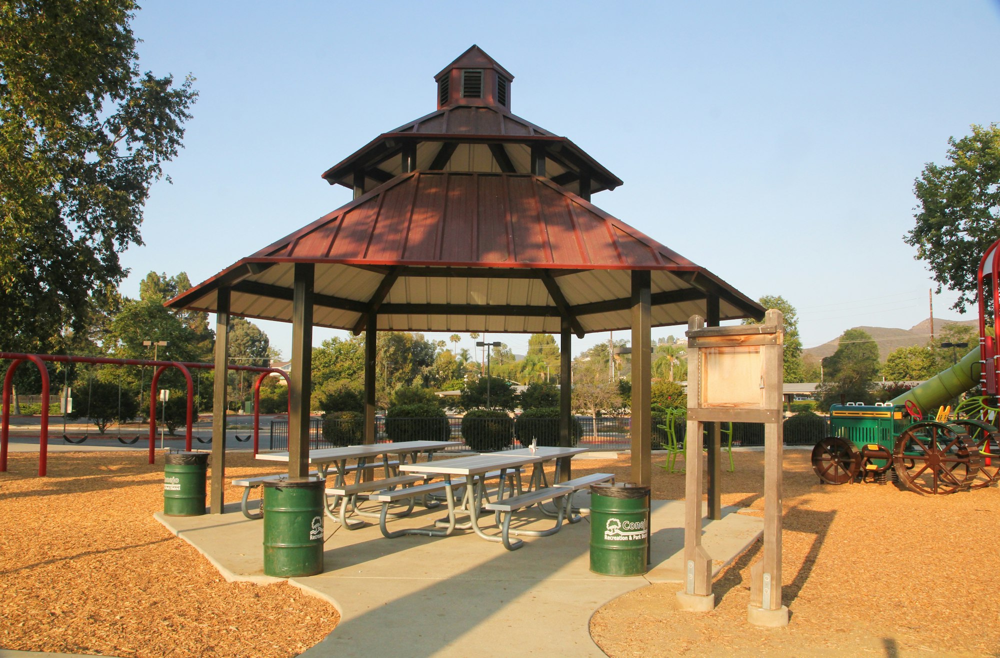A park with a gazebo, picnic tables, trash cans, a playground with swings and slides, set in a scenic area with trees.