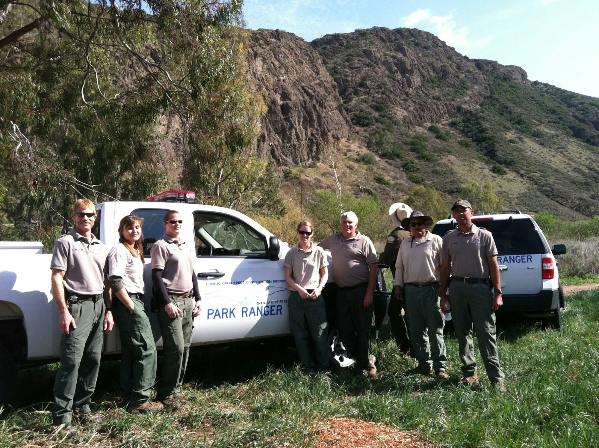 A group of park rangers stands beside park ranger vehicles in a mountainous area.