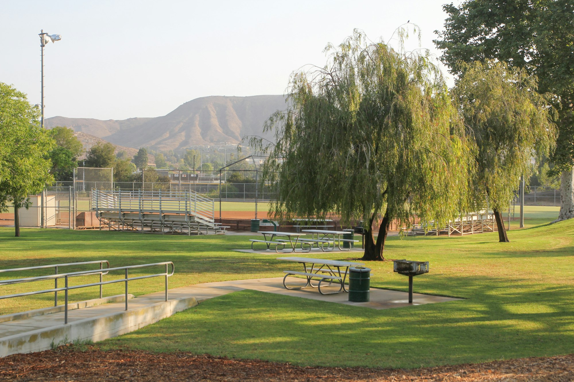 A park with picnic tables, trees, a grill, and a baseball field in the background with bleachers and mountains visible.
