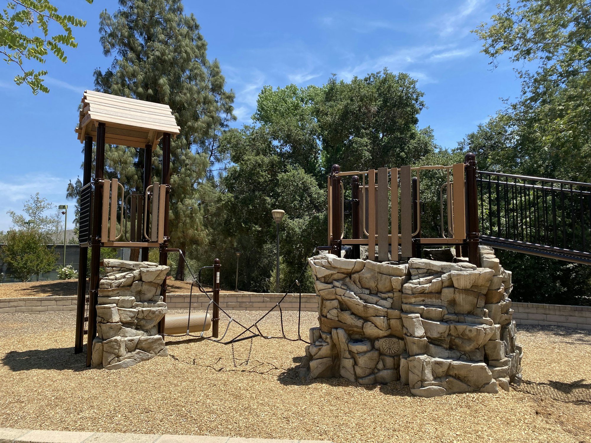 A playground structure with climbing rocks and bridges, surrounded by trees and a woodchip ground.