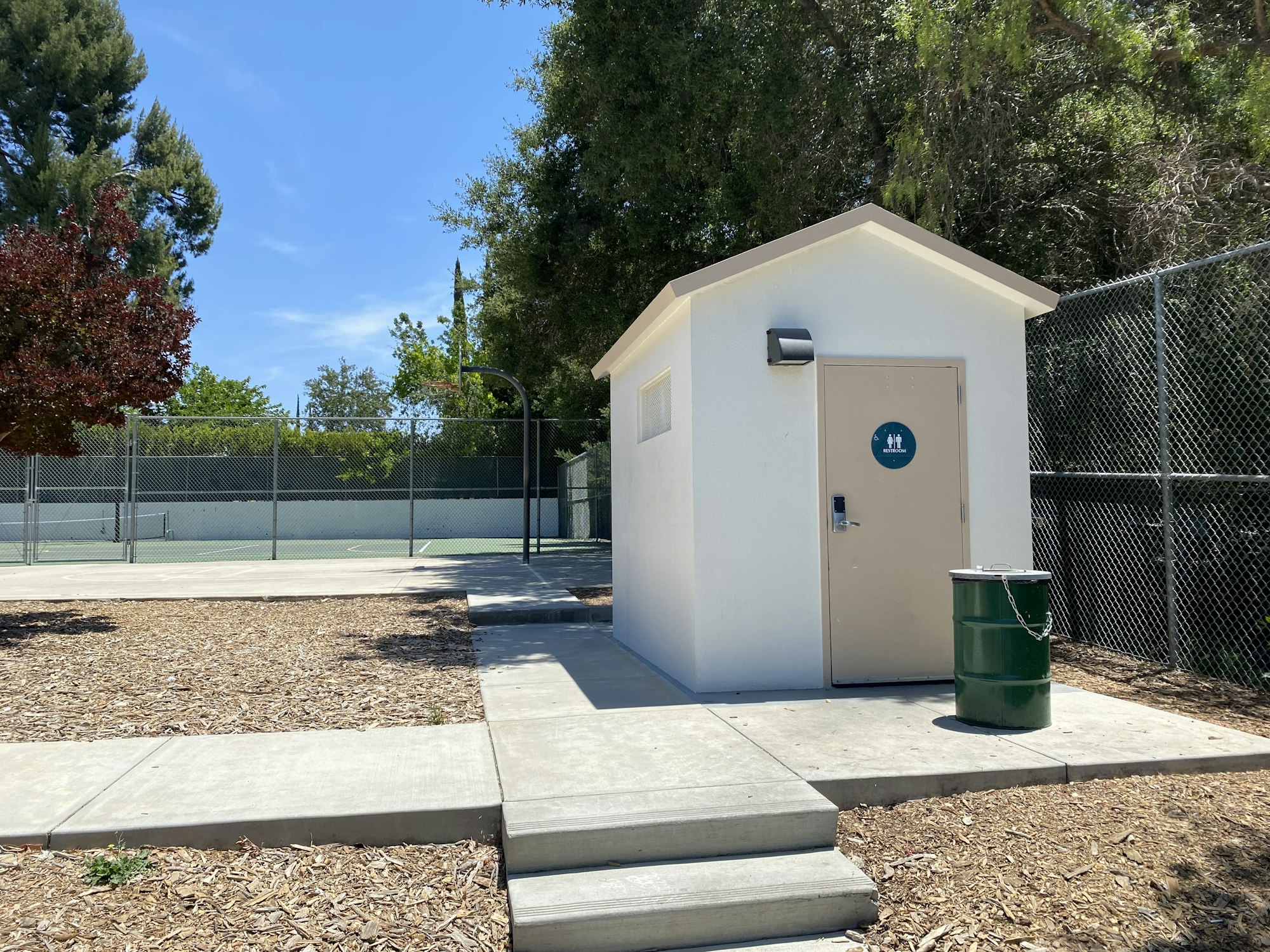 Small building with restroom sign, green trash can chained outside, near a tennis court and trees.